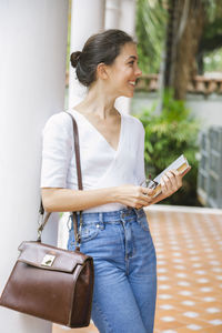 Smiling woman holding book looking away while standing by column