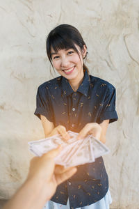 Cropped hand giving paper currency to smiling woman against wall