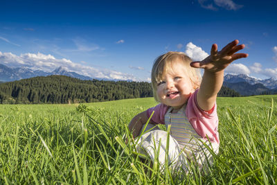 Smiling cute girl gesturing while sitting on grass against sky