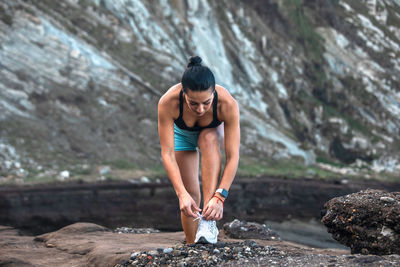 Side view of fit ethnic female athlete tying shoelaces on sneakers while sitting on seashore and preparing for workout