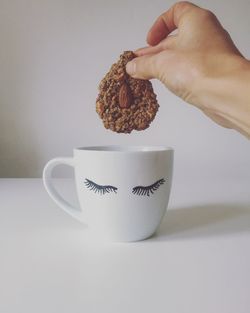 Cropped hand of woman holding cookie over cup on table against white wall