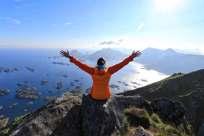 Rear view of woman with arms raised sitting on cliff by sea against sky