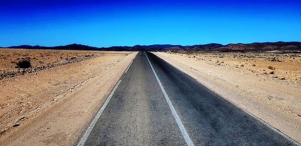 Road leading towards desert against clear blue sky