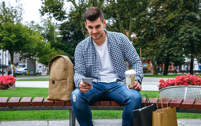 Portrait of young man holding mobile phone and disposable cup while sitting on bench