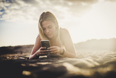 Young woman using mobile phone while lying on sand against sky