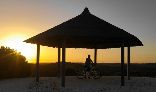 Man with bicycle standing in gazebo against sky during sunset