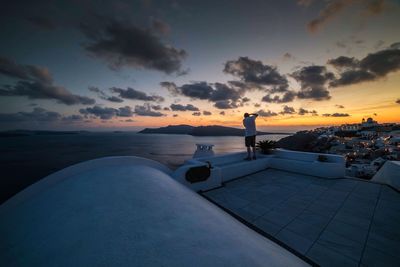 Rear view of man standing on bench by sea against sky during sunset