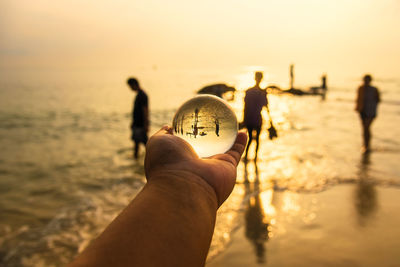 Cropped hand of person holding crystal ball with people in background against sea and sky during sunset