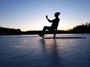 Side view of silhouette man holding wineglass while sitting on sled at frozen lake
