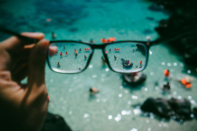 Cropped hand holding eyeglasses against people in sea