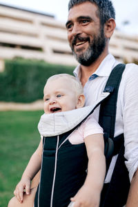 Cheerful adult man in casual clothing with beard looking ahead with toothy smile while carrying laughing infant with sling in park in daylight