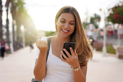 Excited young woman watching her smartphone celebrating outdoor raising her fist up in exultation