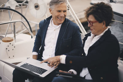 Smiling senior man looking at woman pointing towards laptop while traveling in yacht