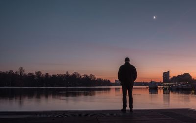 Rear view of silhouette man standing at riverbank against sky during sunset