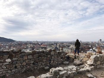 Rear view of person looking at cityscape while standing on old ruin against cloudy sky