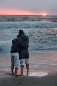 Mother and son standing at beach against sky during sunset