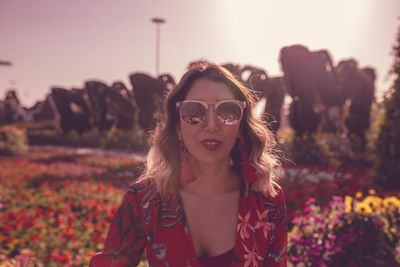 Portrait young woman standing in ornamental garden against sky at dusk