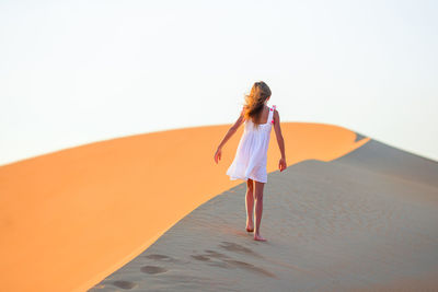 Rear view of girl walking on sand dunes at desert against clear sky