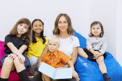 Portrait of mid adult teacher sitting with students against wall at classroom