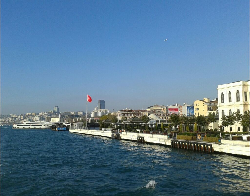 clear sky, building exterior, architecture, built structure, copy space, water, sea, blue, waterfront, flag, city, house, outdoors, day, residential building, river, sky, incidental people, residential structure, rippled