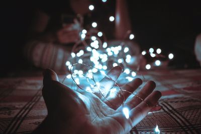 Cropped hand of person holding illuminated string lights on bed