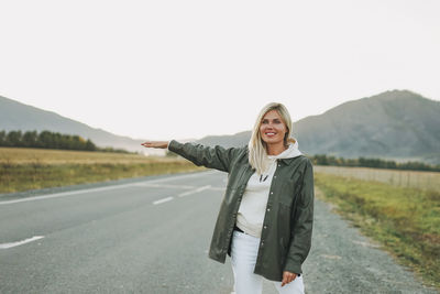 Beautiful smiling blonde young woman traveler in white hoodie on road, trip to mountains