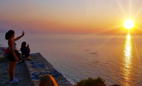 Rear view of people standing by sea against sky during sunset