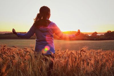 Rear view of woman with arms outstretched standing on wheat field during sunset