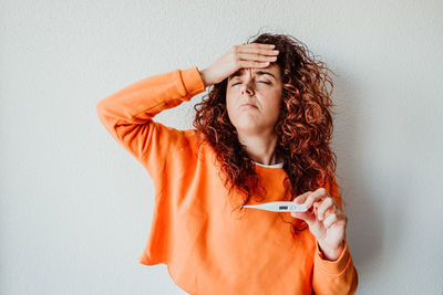 Sick mid adult woman holding thermometer while standing against wall