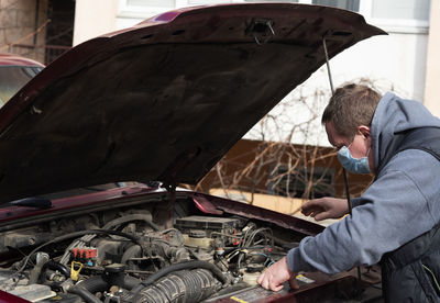 Mechanic in a protective medical mask checks a car engine during a covid-19 coronavirus pandemic