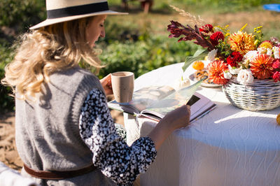 A woman in a hat with a brim flips through a book and drinks a hot drink at a table 