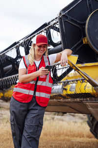 Positive female worker in uniform texting message on cellphone while standing near industrial combine harvester during work in agricultural field
