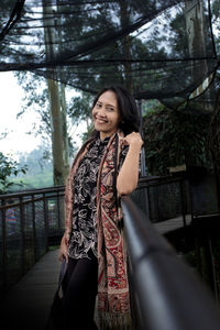 Portrait of smiling woman standing by railing against trees