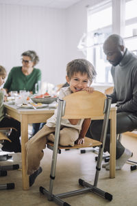 Portrait of smiling boy sitting on chair with teacher at dining table during lunch break