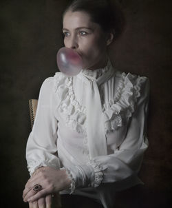 Woman blowing bubble gum while siting on chair against black background