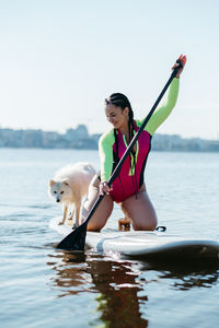 Happy cheerful woman paddleboarding on the city lake at early morning with her dog japanese spitz