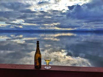 Glass of bottle on table by sea against sky