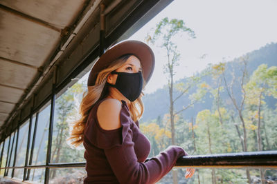 Low angle view of woman standing wearing a mask looking outdoors through a window