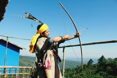 Man holding bow and arrow while standing against sky