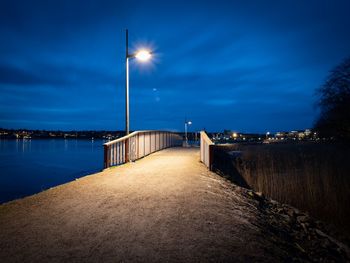 Illuminated street by sea against sky at dusk