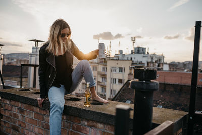 Woman in sunglasses having drink while sitting on retaining wall in city