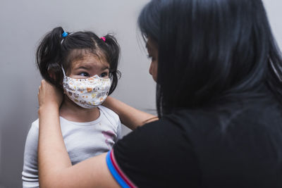 Ethnic loving mother putting protective mask on face of cute little child standing against gray background and looking at each other during coronavirus epidemic