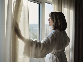 Blond senior woman opening white curtain while looking through window at luxury hotel room