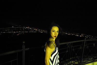 Portrait of young woman standing against illuminated cityscape at night