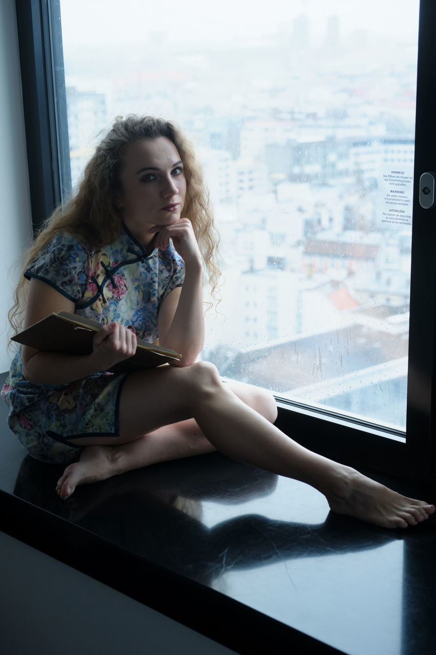 window, one person, women, sitting, indoors, adult, photo shoot, young adult, looking, hairstyle, full length, long hair, architecture, portrait, contemplation, city, day, looking through window, limb, lifestyles, human leg, female, fashion, clothing, blue, person, front view, black, looking away, glass