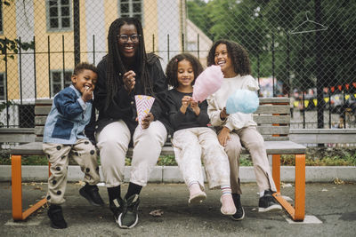 Portrait of happy enjoying with kids eating cotton candies while sitting on bench at amusement park