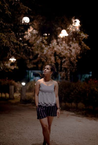 Young woman looking away while standing against trees at night
