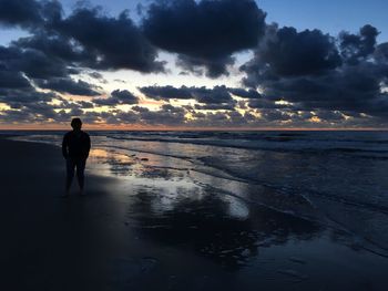 Rear view of silhouette man standing at beach against cloudy sky during sunset