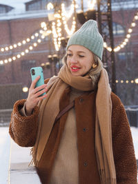 Young stylish smiling blonde woman taking selfie and using smartphone in winter street.