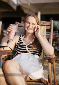 Happy woman talking on mobile phone while sitting on chair with drink